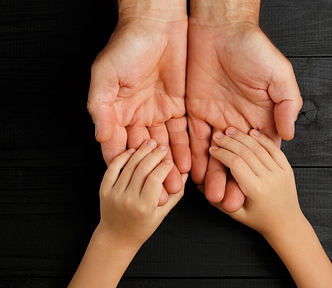 The hands of a child rest gently in the hands of an adult, against a dark wood background.