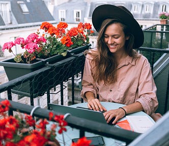 smiling woman sitting on a balcony writing on laptop and surrounded by flowers in window boxes