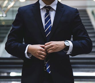A white man in a navy suit adjusts his tie; his face is cropped