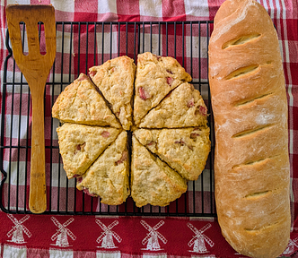 A wooden spatula sits on a metal rack next to freshly baked scones and a loaf of french bread. A red checked cloth is beneath them.