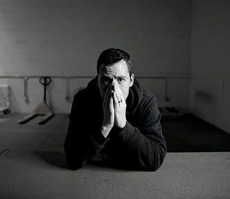 black and white photos of man in empty warehouse wearing black sweatshirt sitting at table with his hands covering his mouth