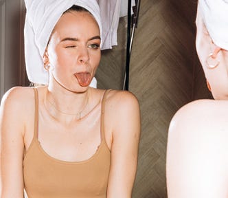 Young woman with hair up in towel and bralette sticking out tongue in mirror