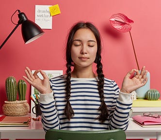 Relaxed Asian woman meditates at workplace, sits in zen pose against desktop, tries to relax after work, isolated over pink background