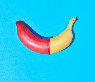 A banana lies on a bright blue background. The left half of the banana is covered with a red condom, creating a sharp contrast with the uncovered yellow right half. The image is brightly lit and has a playful, educational tone, likely symbolizing safe sex practices.