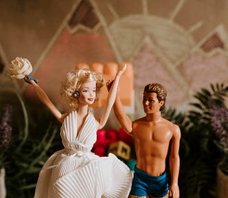 A Barbie doll wearing a white dress and holding a bouquet. She is standing beside a Ken doll wearing only board shorts.