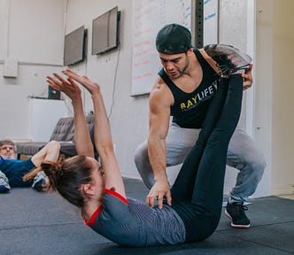 A male trainer at a gym helps a young woman stretch, an older man in the background looks on.