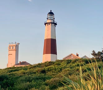 A beige and brown lighthouse, standing on a slightly elevated piece of land. Behind is a clear blue sky and unruly grass on the ground. This photo was taken in Montauk, NY.