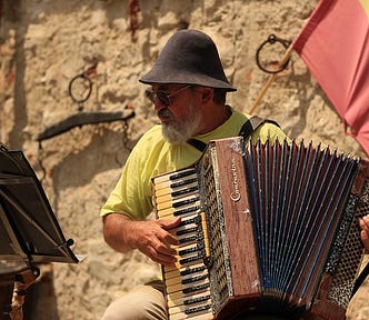 An accordion singer wearing an old-style, ethnic hat while singing in a public place with a flag in the background