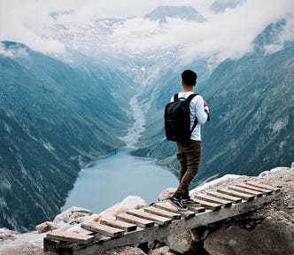 man with backpack walking across wooden bridge looking out toward mountains and river