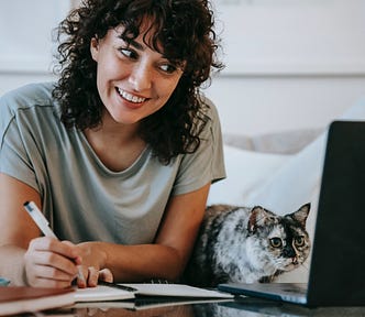 Woman smiling looking at her laptop while writing in a notebook with a cat beside her focused on the laptop.