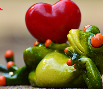 A green and yellow porcelain frog with orange bug eyes laying on its side with a big red heart balanced on his hip