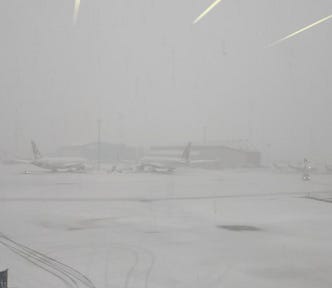 Photo of snow-covered airport tarmac at Boston during a heavy snowstorm, with low visibility obscuring several parked airplanes and terminal buildings in the background