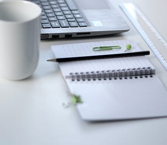 Photo of a laptop, notepad and pencil with paperclip, ruler, and mug on a white table.