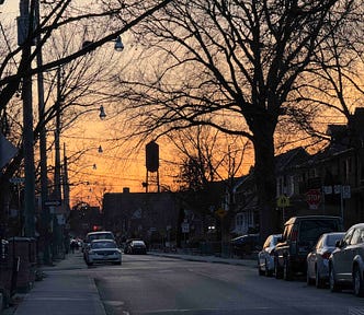 A street scene at dusk with a clear sky transitioning from orange to blue. The street is lined with parked cars on both sides and a few cars driving down the road. Bare trees with no leaves are visible, and streetlights hang from wires above the street. There are houses and buildings along the sides of the street, and a stop sign is visible on the right side of the image. The image captures the calm and quiet atmosphere of a residential neighborhood during sunset.