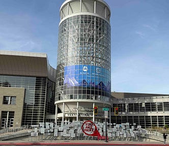 A modern building with a cylindrical glass tower displaying a ‘KubeCon + CloudNativeCon North America 2022’ banner. The intersection in front shows street signs for ‘S TEMPLE’ and ‘200 W.’ Several signs and a large red circle with ‘YOU ARE HERE’ are visible on the ground. Flags are seen on the right side of the image.
