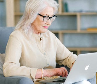 An older woman with shoulder-length gray hit sits in a dusty blue armchair and types on a MacBook computer, which sits on her lap.