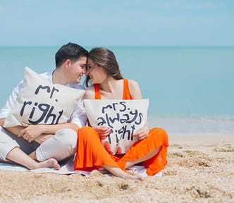 A young couple sitting on a beach with the sea in the background. He is wearing white and holding a cushion that reads Mr right. She is wearing a bright orange dress and holding a cushion that reads Mrs always right.