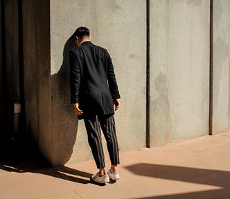 man wearing long-sleeve back top and striped pants leaning head against concrete wall