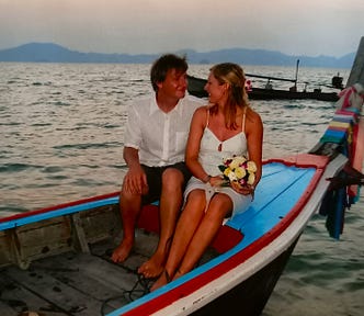 A tanned white couple sit in longtail boat painted blue with red edging. She is wearing a white dress and holding a bouquet and he is wearing a white shirt and pinstriped shorts. A Thai fisherman is holding onto the bow.