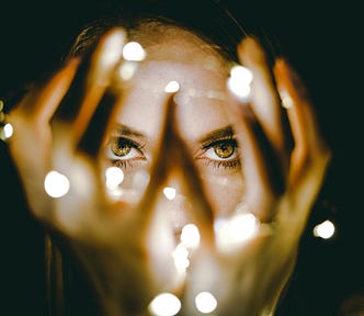 Woman with her hands open around her face with fairy lights wrapped around her hands. Focus is on her hazel eyes.