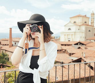 Image of a woman in a hat taking a picture in our direction with a camera with the rooftops of an old, European city behind her.