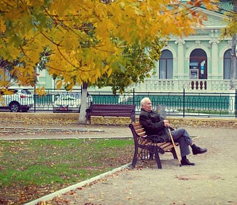 An elderly man sitting on a park bench. He is wearing an overcoat and holding a walking stick. The yellow and gold leaves of two trees are visible in shot, and fallen leaves litter the ground. There are no other people visible in the park.