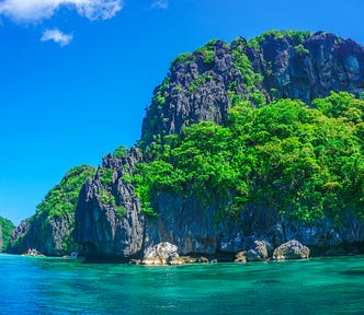 The rocky cliff of a tropical island covered with low lush green bushes. Below is crystal clear turquoise water.