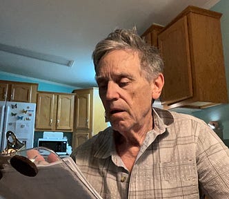 An older man with gray hair wearing a plaid shirt is standing in a kitchen, looking thoughtfully at papers held on a clipboard.