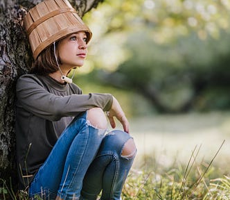 A young girl, perhaps nine years old, sitting against the trunk of a large tree. She has holes in the knees of her jeans, and an upturned wooden bucket on her head, the handle of which hangs like a strap under her chin.