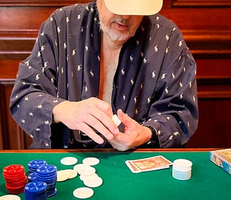 An elderly man playing poker in a bathrobe and a baseball cap, actively counting his chips.