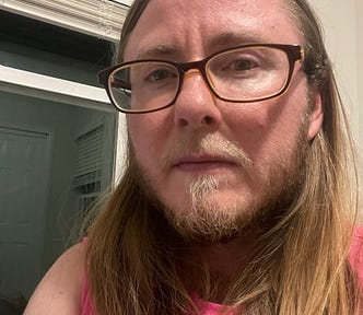 The author is wearing brown framed glasses and a pink shirt. He has a defiant look on his face. He’s standing in front of a window. It’s dark outside.
