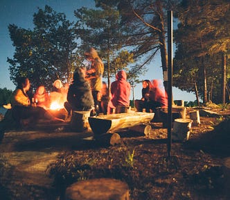 group of people sit around a campfire talking in a forest at dusk