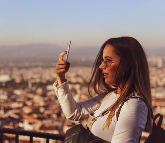 Photo Of Woman Using Mobile Phone. she’s set against a cityscape in the background. she’s probably taking a picture. she looks happy. i picked this photo because she’s on her phone, potentially making money, while out travelling.