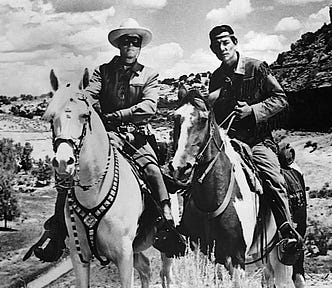 Black-and-white photo of the Lone Ranger, a man wearing a mask covering his eyes and a cowboy hat, sitting on a white horse (left) and Tonto, a Native American, sitting on a multi-color horse (right) set against a rugged background.