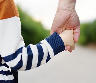 A child holds the hand of an adult while they walk down a wooded path.
