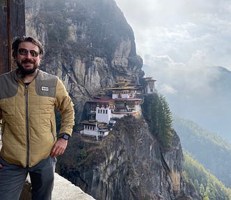 The author, in the foreground, wearing a beige colored warm weather jacket. Tigers Nest Monastery buildings, white with brown roofs, are in the background, clinging to the side of a steep rocky mountain. More clouds and mountains are visible in the distance.