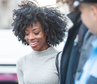A Black woman in a cream turtleneck is looking down and smiling widely as a white police officer looks at her.