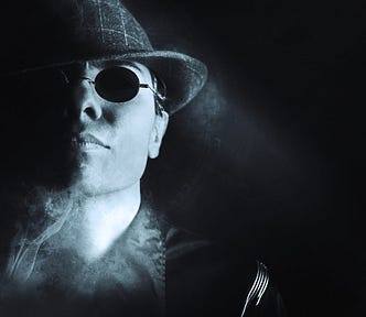 Man, looks mysterious /menacing, wears trilby hat and dark glasses