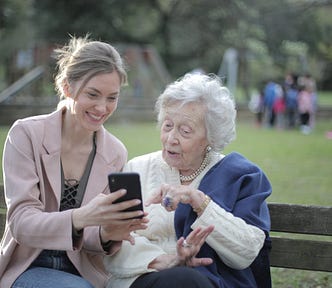 A middle-aged woman and an elderly woman, both smartly dressed, sitting on a park bench. The older woman is showing great interest in the phone held by the younger.