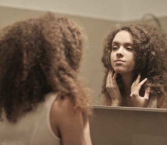 A young woman with tightly curled brown hair looks at herself in a large bathroom mirror.