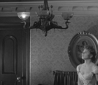 A black and white still from the 1944 film Gaslight. Paula, played by Ingrid Bergman, stares up with a haunted expression at a flickering gaslight chandelier in her home. Just one of its light is on.