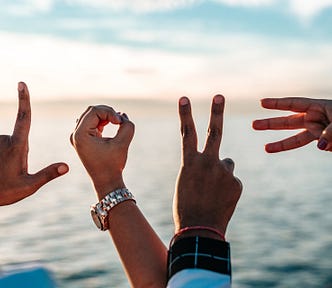 Photo of four hands, each spelling out a letter for the word “LOVE” The background is a blurry seascape.