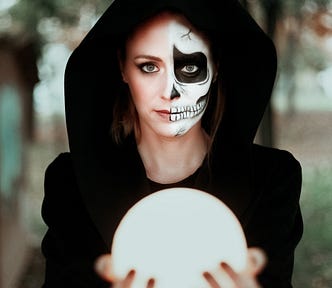 Woman in a hooded robe holding out a crystal ball, her face half made up to look like a skull