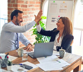 A businesswoman and businessman are high-fiving across a table from each other, both with smiles on their faces