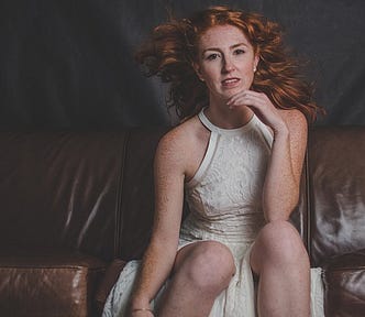 Attractive woman, long red hair, cream party dress, bare arms, seated on leather couch