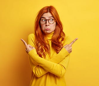 A woman with long ginger hair has her arms crossed with index fingers pointing in different directions. She looks confused. She is wearing yellow against a yellow background.