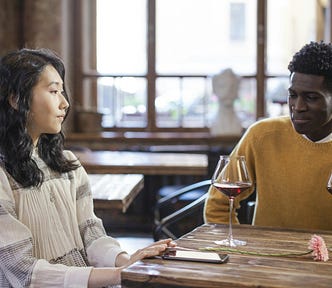 Asian woman and black man sitting at a table and each drinking a cup of wine