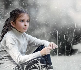 A pre-teen girl with dark hair and piercing green eyes sitting in the snow beside a fir tree. She is wearing black leggings and a white turtleneck sweater.