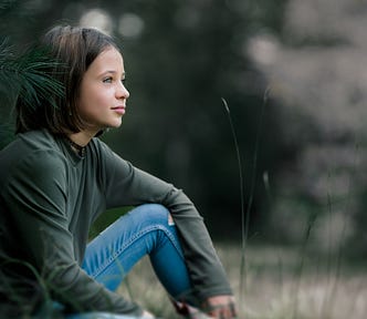 A young girl, perhaps nine years old, with intense, watchful eyes, sitting on grass beside a conifer. She has holes in the knees of her jeans.