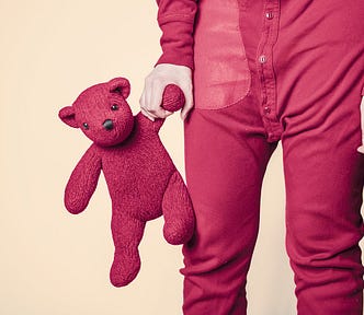 Image with a yellowish background, and to the right of the image stands a grown person wearing dark red and holding a teddy in the same color.